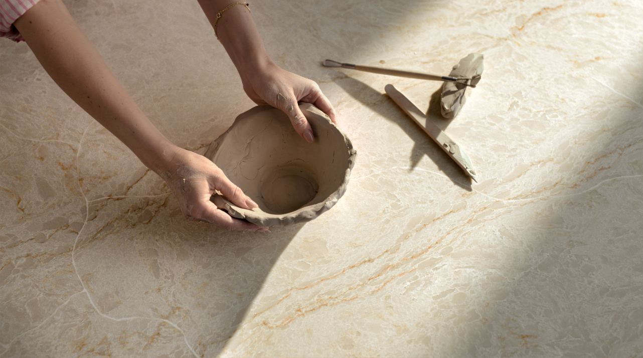 Hands shaping clay pottery on a surface, with carving tools nearby in sunlight.